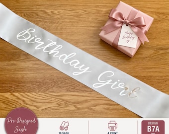 May include: A silver satin sash featuring the white cursive text Birthday Girl, accompanied by a square pink gift box tied with a matching satin ribbon and a small white tag labeled Happy Birthday with a heart icon.