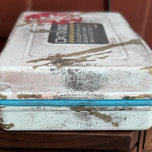 May include: A rectangular white storage case with a distressed finish featuring gold paint splatters and a bright blue rubber seal. The top surface displays a black rectangular label with yellow hazard stripes and white text, alongside a red circular logo.