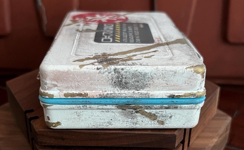 May include: A rectangular white storage case with a distressed finish featuring gold paint splatters and a bright blue rubber seal. The top surface displays a black rectangular label with yellow hazard stripes and white text, alongside a red circular logo.