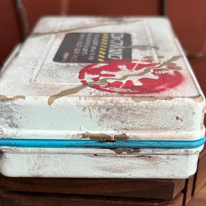 May include: A rectangular white metal storage box with a distressed finish, featuring a bright blue trim along the center seam. The lid displays a red circular graphic with a jagged gold line and a black rectangular label with white text.