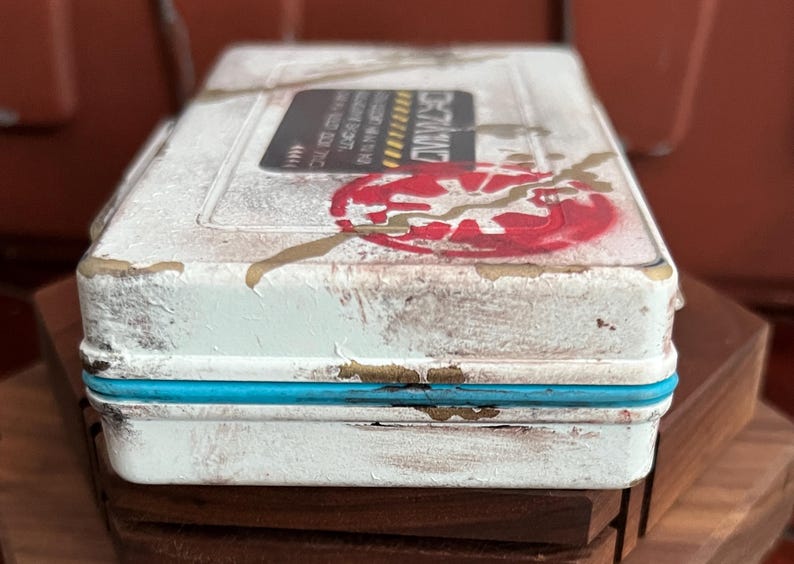 May include: A rectangular white metal storage box with a distressed finish, featuring a bright blue trim along the center seam. The lid displays a red circular graphic with a jagged gold line and a black rectangular label with white text.