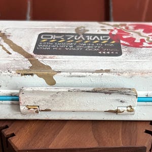 May include: A rectangular white storage case with a distressed finish, featuring gold paint splatters and a bright blue trim. A black rectangular label on the lid displays stylized text and a red circular emblem with a white symbol in the center.