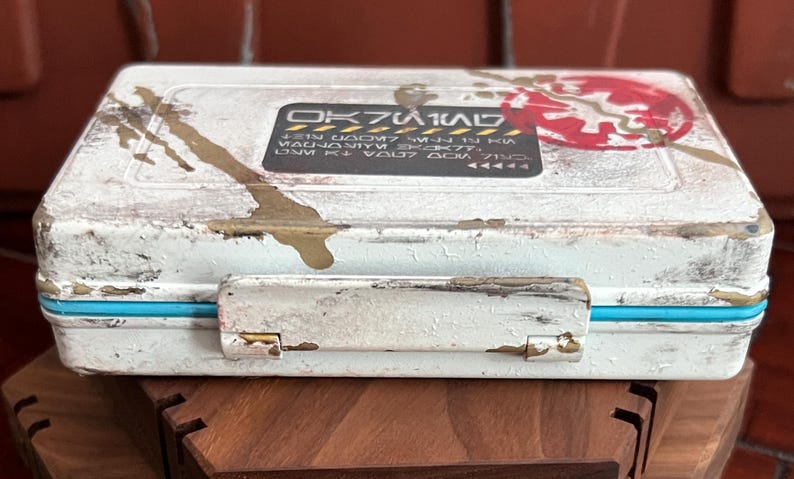 May include: A rectangular white storage case with a distressed finish, featuring gold paint splatters and a bright blue trim. A black rectangular label on the lid displays stylized text and a red circular emblem with a white symbol in the center.