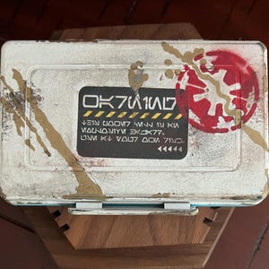 May include: A rectangular white metal storage box with a weathered finish, featuring gold paint splatters and a red circular emblem. A central black label displays stylized text reading OK7W1N7, followed by additional lines of fictional script.