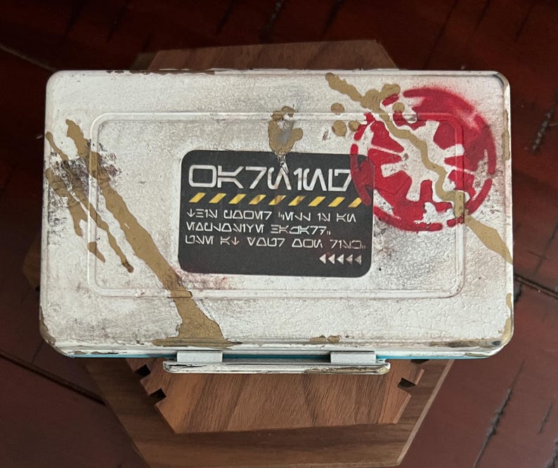 May include: A rectangular white metal storage box with a weathered finish, featuring gold paint splatters and a red circular emblem. A central black label displays stylized text reading OK7W1N7, followed by additional lines of fictional script.