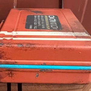 May include: A rectangular orange metal storage case with a distressed finish, featuring a bright turquoise trim along the center seam and two horizontal white stripes. A black rectangular label on the top displays white text and yellow accents.