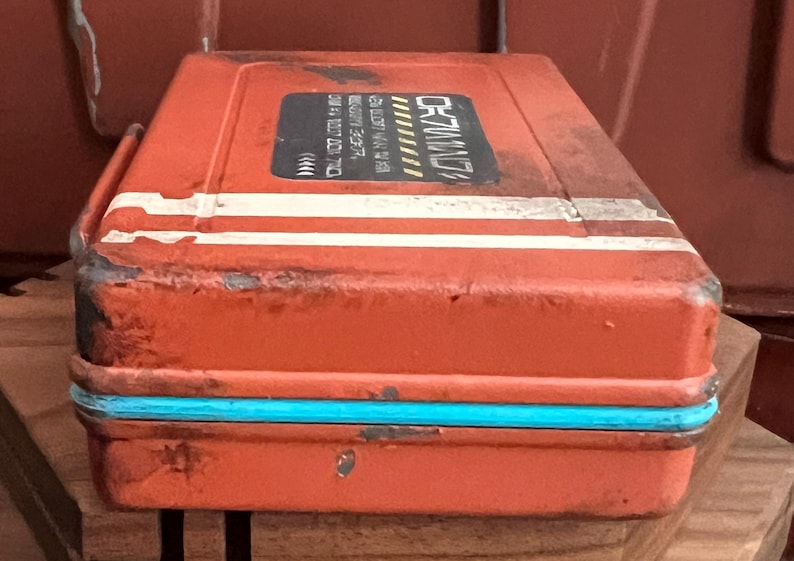 May include: A rectangular orange metal storage case with a distressed finish, featuring a bright turquoise trim along the center seam and two horizontal white stripes. A black rectangular label on the top displays white text and yellow accents.