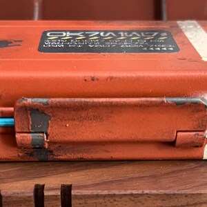 May include: A rectangular orange storage case with a distressed finish, featuring white stripes on the right side and a bright blue trim along the center seam. A black rectangular label with white text and yellow hazard stripes is on the top lid.
