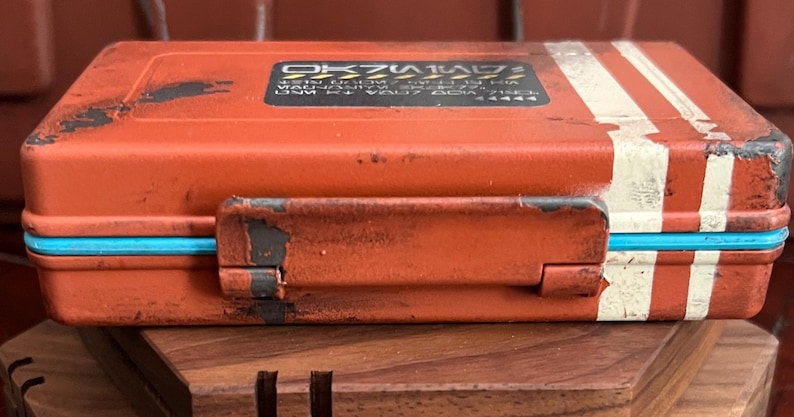 May include: A rectangular orange storage case with a distressed finish, featuring white stripes on the right side and a bright blue trim along the center seam. A black rectangular label with white text and yellow hazard stripes is on the top lid.