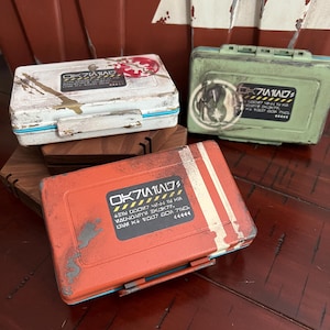 May include: Three rectangular storage cases in white, green, and orange, each featuring a weathered finish, a hinged lid, and a central label with black text and yellow hazard stripes. The cases display unique circular emblems and sci-fi style lettering.