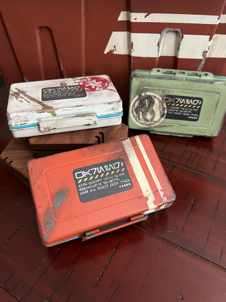 May include: Three rectangular storage cases in white, green, and orange, each featuring a weathered finish, a hinged lid, and a central label with black text and yellow hazard stripes. The cases display unique circular emblems and sci-fi style lettering.
