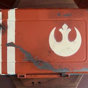 May include: A rectangular orange storage box with a distressed finish featuring two vertical white stripes on the left side and a white Rebel Alliance symbol on the right. The surface shows signs of wear with dark gray paint chips and scratches.