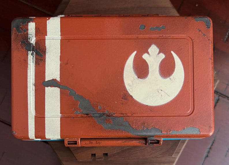 May include: A rectangular orange storage box with a distressed finish featuring two vertical white stripes on the left side and a white Rebel Alliance symbol on the right. The surface shows signs of wear with dark gray paint chips and scratches.