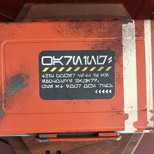 May include: A rectangular orange storage box with a distressed, weathered finish and two vertical white stripes on the right side. A central black rectangular label features stylized white text and yellow hazard-style diagonal stripes.