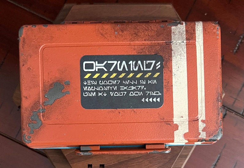 May include: A rectangular orange storage box with a distressed, weathered finish and two vertical white stripes on the right side. A central black rectangular label features stylized white text and yellow hazard-style diagonal stripes.