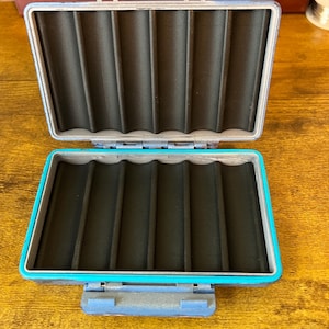 May include: A rectangular, open storage case featuring a bright turquoise outer frame and a dark grey, hinged lid. The interior is lined with black foam that has six vertical, rounded channels designed for holding slender items securely in place.