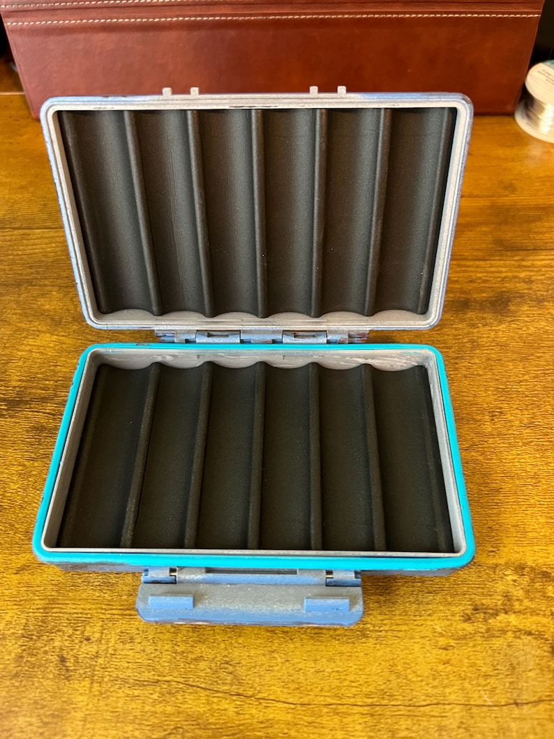 May include: A rectangular, open storage case featuring a bright turquoise outer frame and a dark grey, hinged lid. The interior is lined with black foam that has six vertical, rounded channels designed for holding slender items securely in place.