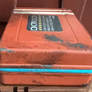 May include: A rectangular, weathered orange storage case with a distressed finish, featuring a bright turquoise horizontal band along the center. A black rectangular label on the top displays white text and yellow hazard stripes, showing the word CH7.