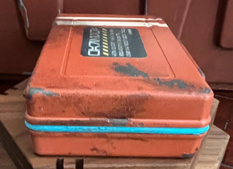 May include: A rectangular, weathered orange storage case with a distressed finish, featuring a bright turquoise horizontal band along the center. A black rectangular label on the top displays white text and yellow hazard stripes, showing the word CH7.