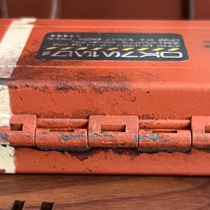 May include: A rectangular orange storage case with a weathered finish, featuring two vertical white stripes on the left side and a bright blue trim along the center seam. A black rectangular label with white text is centered on the top lid.