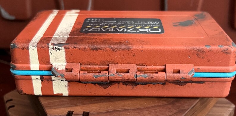 May include: A rectangular orange storage case with a weathered finish, featuring two vertical white stripes on the left side and a bright blue trim along the center seam. A black rectangular label with white text is centered on the top lid.