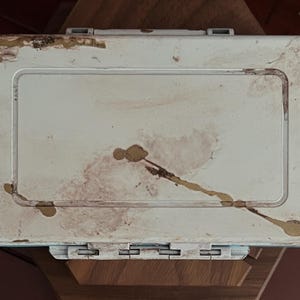 May include: A rectangular, off-white plastic storage case featuring a hinged lid with a central recessed panel. The exterior surface shows visible signs of wear, including brown stains, scuffs, and patches of peeling material along the edges.
