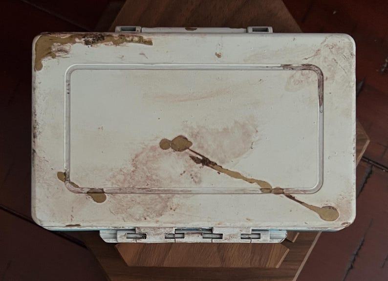 May include: A rectangular, off-white plastic storage case featuring a hinged lid with a central recessed panel. The exterior surface shows visible signs of wear, including brown stains, scuffs, and patches of peeling material along the edges.