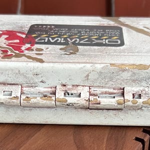 May include: A rectangular white storage case with a distressed finish, featuring a red circular emblem on the left and a black rectangular label with white text on the top. The case has a bright blue trim along the center seam and metal hinges.
