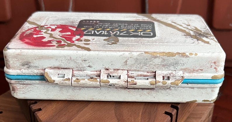 May include: A rectangular white storage case with a distressed finish, featuring a red circular emblem on the left and a black rectangular label with white text on the top. The case has a bright blue trim along the center seam and metal hinges.