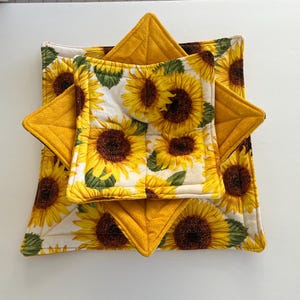 May include: A set of quilted bowl cozies and pot holders. The bowl cozies and pot holders feature a bright yellow sunflower pattern on a white background. The pot holders are a solid yellow color. The set is designed to protect hands and surfaces from heat.
