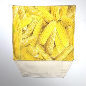 May include: A rectangular fabric pouch featuring a vibrant, repeating photographic print of numerous yellow corn cobs. The top section displays the detailed corn pattern, while the bottom section consists of a plain, off-white canvas fabric.
