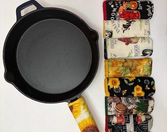 May include: A black cast iron skillet with a yellow sunflower handle cover. Next to the skillet are several fabric handle covers with various patterns, including apples, tomatoes, sunflowers, and farm-themed designs.