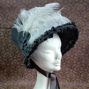 May include: A dark gray bonnet with a ruffled brim and white feathers. The hat is displayed on a white mannequin head with a dark gray ribbon tied around the neck. The hat is a vintage style.