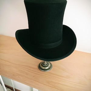 Può includere: Un cappello a cilindro nero, un classico copricapo formale, con una corona cilindrica alta e un'ampia tesa. Il cappello è realizzato in un materiale scuro ed è esposto su un supporto decorativo color argento. Un accessorio senza tempo.