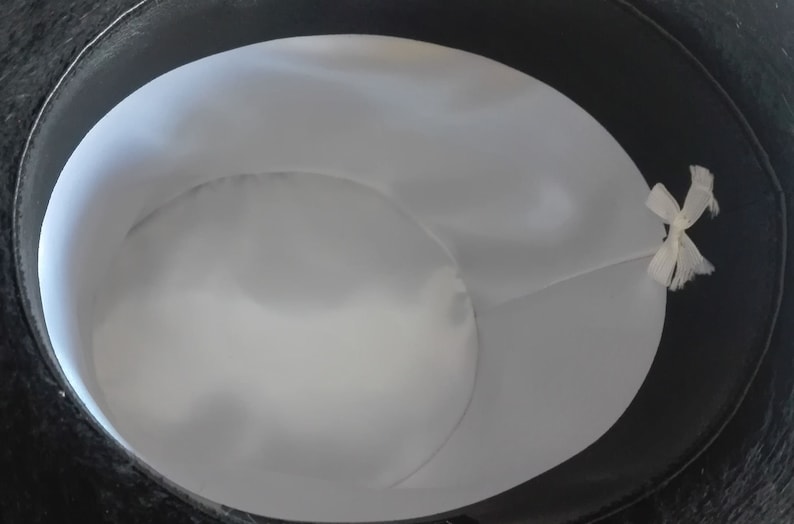 Puede incluir: Un sombrero de copa negro con forro blanco y una cinta blanca atada en el interior.