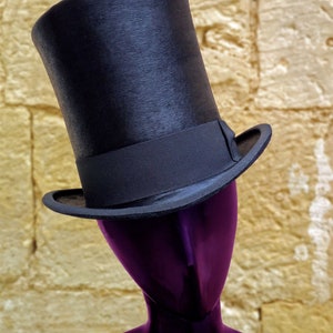 Puede incluir: Un sombrero de copa negro con una banda negra se sienta sobre una cabeza de maniqu&iacute; p&uacute;rpura. El maniqu&iacute; est&aacute; contra una pared amarilla texturizada.