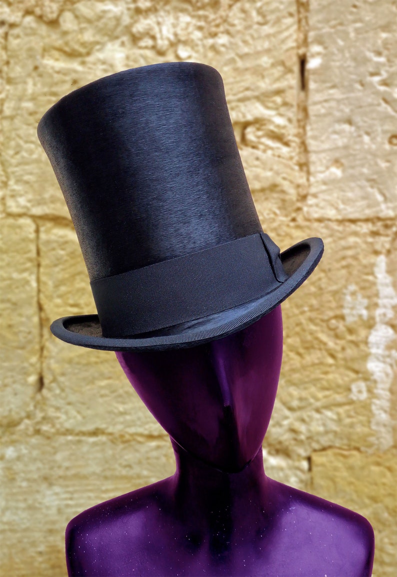 Puede incluir: Un sombrero de copa negro con una banda negra se sienta sobre una cabeza de maniqu&iacute; p&uacute;rpura. El maniqu&iacute; est&aacute; contra una pared amarilla texturizada.