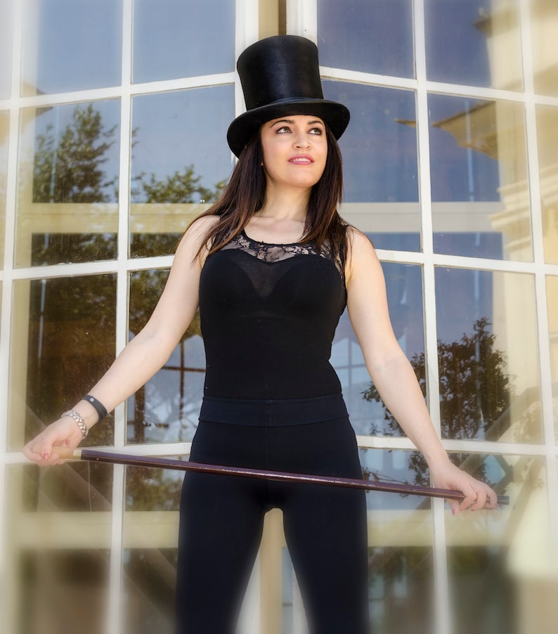 Puede incluir: Una mujer con un top negro y pantalones negros con un sombrero de copa negro. Est&aacute; sosteniendo un bast&oacute;n de madera.