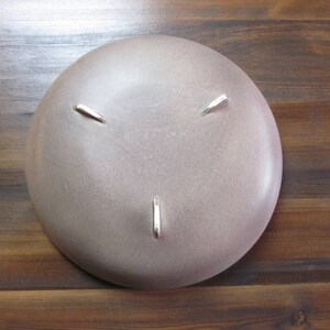 May include: A light brown, round ceramic bowl with three silver-colored feet. The bowl has a smooth surface and a slightly textured appearance. The bowl is sitting on a wooden surface.