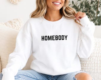 Puede incluir: Una sudadera blanca de manga larga con cuello redondo que presenta la palabra HOMEBODY impresa en letras mayúsculas negras en el pecho. La prenda tiene cuello, puños y dobladillo acanalados, ofreciendo un aspecto limpio y sencillo.