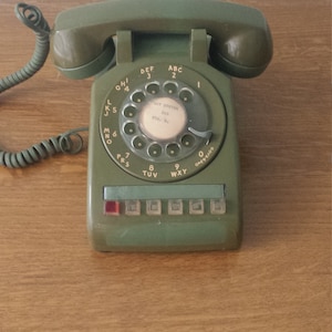 Può includere: Un telefono a disco verde con un disco rotatorio nero e un quadrante bianco. Il telefono ha un filo e una cornetta. Il quadrante ha le lettere ABC, DEF, GHI, JKL, MNO, PRS, TUV, WXY e 0. Il quadrante ha anche il testo "AST SYSTEM 203 STA. 82".