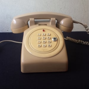 Op de afbeelding: Een beige draaitelefoon met een toetsenbord. De telefoon heeft een rood lampje bovenop en een snoer dat aan de basis is bevestigd.