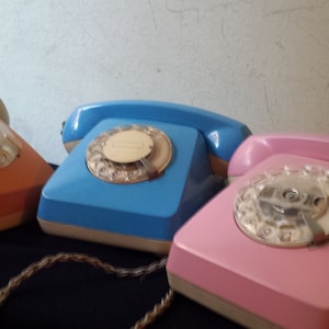 Könnte beinhalten: Drei Vintage-Telefone mit Wählscheibe in Orange, Blau und Rosa. Jedes Telefon hat eine kreisförmige Wählscheibe und einen Hörer. Die Telefone sind in einer Reihe angeordnet, das orangefarbene Telefon links, das blaue in der Mitte und das rosa rechts.