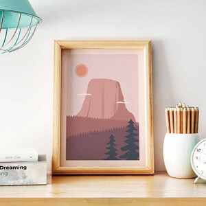 May include: A framed print of a minimalist illustration of a mountain range with a pink sky and a sun. The mountain range is in the center of the print and is surrounded by trees. The print is in a light wood frame.