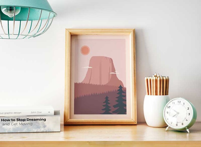 May include: A framed print of a minimalist illustration of a mountain range with a pink sky and a sun. The mountain range is in the center of the print and is surrounded by trees. The print is in a light wood frame.