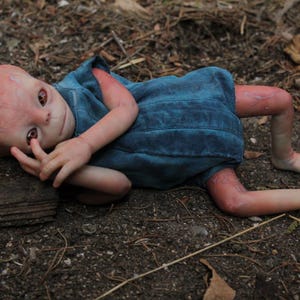 Puede incluir: Una muñeca realista de bebé alienígena con piel rosada-marrón, ojos marrones grandes y un atuendo de mezclilla azul. La muñeca está tumbada en el suelo, rodeada de hojas y tierra, con las extremidades extendidas.