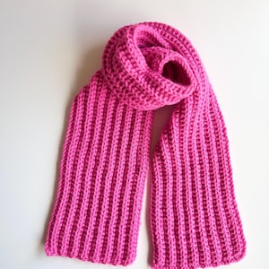 May include: A vibrant pink, ribbed-knit scarf. The scarf is made of a soft, thick yarn and is folded to display its length and texture. The bright color and chunky knit suggest warmth and style.