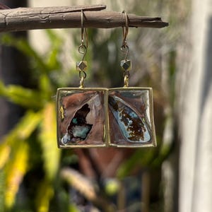 Può includere: Orecchini quadrati con un design color rame e turchese, incorniciati in metallo dorato. Gli orecchini pendono da ganci per orecchie color oro e sono sospesi a un piccolo ramo su uno sfondo verde sfocato.
