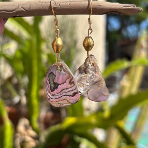 May include: Handmade earrings with iridescent abalone shell pendants in pink, purple, and green hues. The earrings have gold-coloured metal hooks and a gold bead accent. They are displayed on a natural wooden stick against a blurred green background.