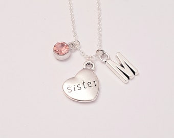 Pode incluir: Um colar de prata com um pingente em forma de coração que diz "irmã" e um pingente com a letra "M". O colar também tem um pequeno pingente de cristal rosa.